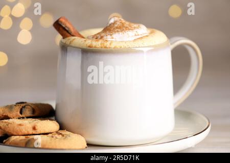 Eine Tasse leckeren Eierpunsch mit Zimt und Keksen auf dem Tisch vor festlichen Lichtern, Nahaufnahme Stockfoto