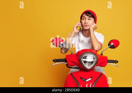 Der Lieferjunge im weißen T-Shirt sitzt auf einem roten Roller vor gelbem Hintergrund, hält das Telefon am Ohr, schnelles Lieferkonzept, Kopierraum Stockfoto