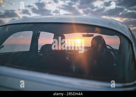 Bringen Sie mich überall hin... Rückblick auf ein Paar, das während einer Autofahrt anhält, um den Sonnenuntergang zu beobachten. Stockfoto