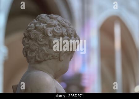 Nahaufnahme der Marmorstatue Rückseite des Kopfes eines kleinen Kindes, Engelchen, mit Locken-Haardetails. Dom-Wand mit Prismenlichteffekt im Hintergrund. Stockfoto