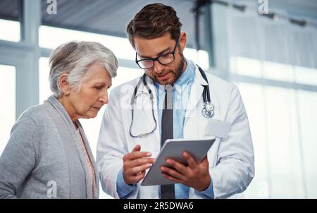 Hier liegt das Problem... ein Arzt, der etwas auf einem digitalen Tablet mit einem leitenden Patienten in einer Klinik diskutiert. Stockfoto