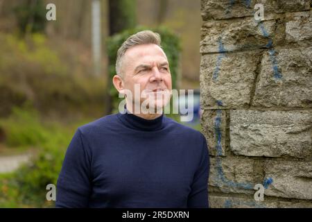 Ein Mann mit grauhaarigen Haaren im blauen Pullover steht neben einer Wand aus großen Steinen und sieht seitlich verärgert aus Stockfoto