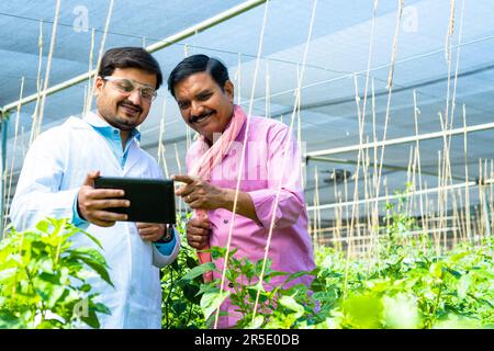 Indischer glücklicher Agrarwissenschaftler mit Farmer, der ein Tablet über das Pflanzenwachstum im Gewächshaus überprüft - Konzept von Expertenhilfe, Anleitung und Biotechnologie Stockfoto