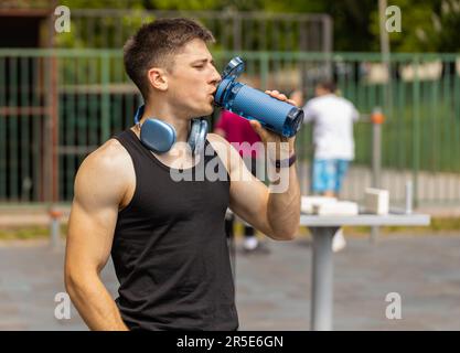 Dehydrierter durstischer sportlicher Kaukasier Muskelmann, der Wasser aus der Flasche trinkt. Ein müder, erschöpfter Kerl auf dem Spielplatz. Sport, Gesundheit, Fitness Routine, Training. Läufer nach schwierigem Joggen im Freien Stockfoto