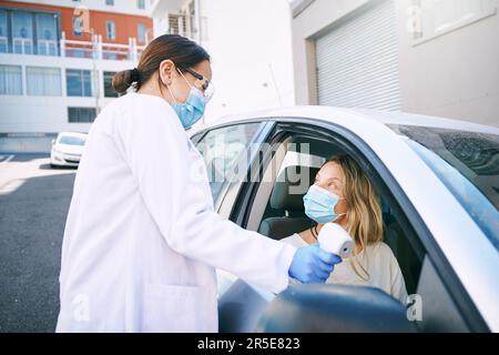 Achten Sie darauf, dass die Temperatur auf der Markierung liegt. Eine maskierte junge Frau, die sich von einem Arzt die Temperatur messen lässt, während sie in ihrem Auto sitzt. Stockfoto