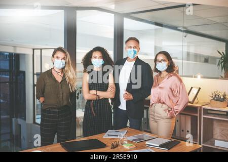 Nichts kann uns davon abhalten, Erfolg zu haben, selbst Covid. Eine vielfältige Gruppe von Geschäftsleuten, die im Büro zusammenstehen und Gesichtsmasken tragen. Stockfoto