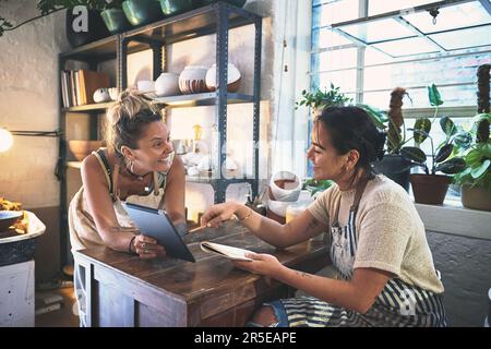 Eine Kombination aus künstlerischem Talent und Geschäftssinn für eine erfolgreiche Kombination. Zwei junge Frauen, die während eines Meetings in einem Töpferstudio ein digitales Tablet benutzen. Stockfoto