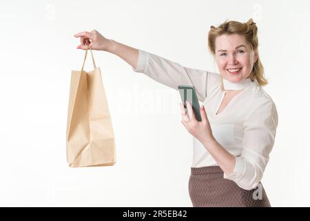 Fröhliche Käuferin, die Einwegpapiertaschen in ausgestreckter Hand hält und mit dem Smartphone bargeldlos bezahlt. Lächelnde Frau Stockfoto