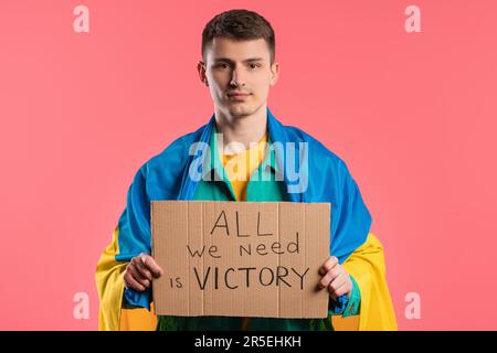 Ein ukrainischer Mann mit Pappe alles was wir brauchen ist ein Sieg auf pinkfarbenem Hintergrund. Stockfoto