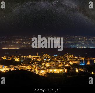 Blick auf die Stadt Lentini bei Nacht in Richtung Bergstraße zum Meer und Ätna-Vulkan (Siracusa, Sizilien, Italien) und die Milchstraße oben. Stockfoto