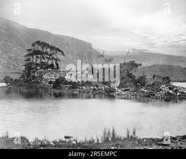 Ein Blick aus dem späten 19. Jahrhundert auf die Eremitage-Ruinen in Gougane Barra aus dem Jahr 1700, als sich ein Priester namens Denis O'Mahony auf die Insel an einem malerischen Talsee in den Shehy Mountains der Grafschaft Cork, Irland, zurückzog. Der Name Guagán Barra stammt aus der Heiligen Finnbarr (oder Barra), der angeblich im sechsten Jahrhundert ein Kloster auf der Insel im See gebaut hat. Stockfoto