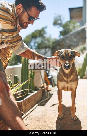 Wer will spazieren gehen? Ein fröhlicher junger Mann, der tagsüber seinen Hund vor seinem Haus streichelt. Stockfoto
