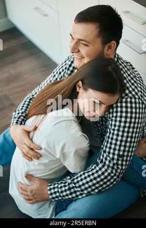 Lächelnde junge Ehepartner sitzen, umarmen sich sanft, ein Stockwerk in der Küche Stockfoto