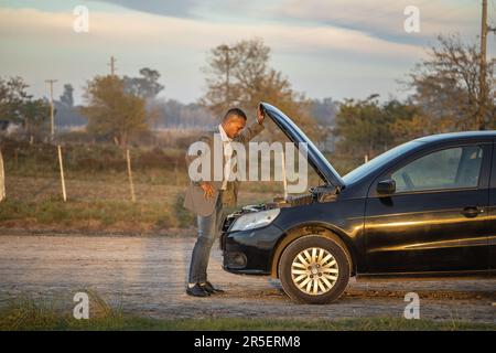 Ein junger Latino in einem Anzug, der den Motor seines kaputten Autos am Straßenrand ansieht. Stockfoto