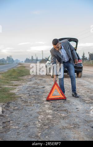 Ein junger Latino-Mann, der ein Warndreieck hinter einem kaputten Auto am Straßenrand platziert. Stockfoto