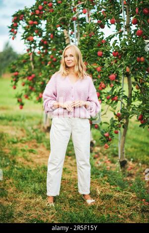 Außenporträt eines romantischen jungen weiblichen Models, das sich in einem Apfelgarten posiert, eine lila Strickjacke und weiße Hosen trägt Stockfoto