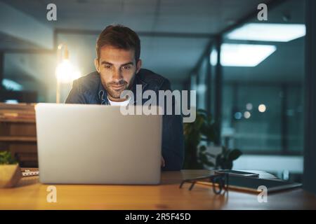 Fokussiert und fleißig. Ein gutaussehender junger Geschäftsmann, der spät in der Nacht an seinem Computer in einem modernen Büro arbeitet. Stockfoto