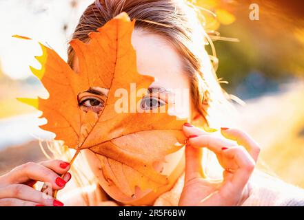 Schöne junge Frau bedeckt Gesicht mit Blatt. Nahaufnahme Porträt von schönen Mädchen Gesicht in der Nähe bunte Herbstblätter. Nette Frau hält Herbstblätter Stockfoto