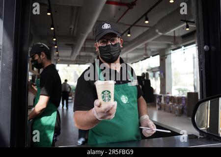 Starbucks Mitarbeiter geben Befehle mit fröhlichem Gesichtsausdruck und tragen Gesichtsmasken am Drive-in. Eisgekühlter weißer Mokka-Kaffee. Stockfoto