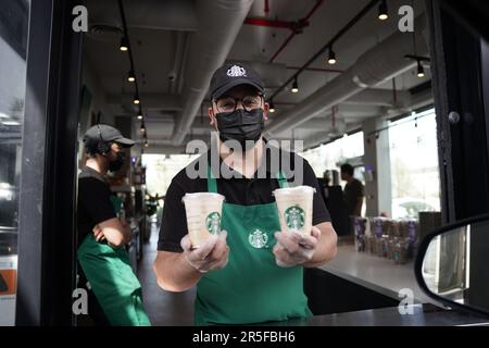 Starbucks Mitarbeiter geben Befehle mit fröhlichem Gesichtsausdruck und tragen Gesichtsmasken am Drive-in. Eisgekühlter weißer Mokka-Kaffee. Stockfoto