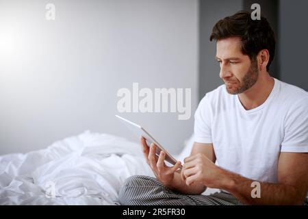 Er meint es ernst, eine Lösung für seine Schlaflosigkeit zu finden. Ein gutaussehender Mann mittleren Alters, der sein digitales Tablet benutzt, während er sich in seinem Schlafzimmer entspannt. Stockfoto
