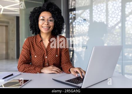 Junge, wunderschöne afroamerikanische Frau, die in einem modernen Büro arbeitet, eine lächelnde Geschäftsfrau, die mit einem Laptop vor der Kamera auf die Arbeit schaut. Stockfoto