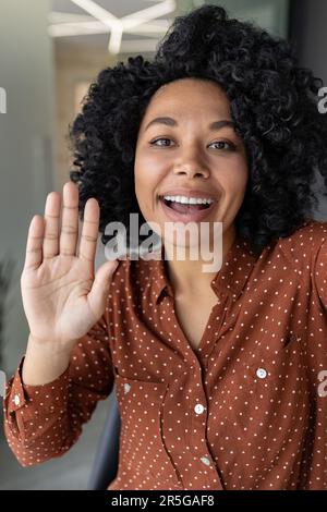 Vertikale Aufnahme, junge, wunderschöne afroamerikanische Geschäftsfrau mit lockigem Haar, die bei Videogesprächen spricht, vor der Smartphone-Kamera winkt, mit der Remote-Kommunikations-App. Stockfoto