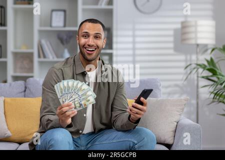 Porträt eines jungen, glücklichen lateinamerikanischen Mannes, der sitzt, Bargeld in der Hand hält, ein Telefon in der Hand hält, auf die Kamera zeigt, lächelt, glücklich. Stockfoto