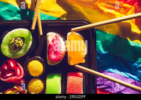 Sushi-Süßigkeiten LGBT-Schwulenparty. Sushi in einer Schachtel auf hellen, mehrfarbigen Regenbogenhintergründen von oben. Hölzerne Sushi-Stäbchen Essstäbchen Asiatisches Japan Stockfoto