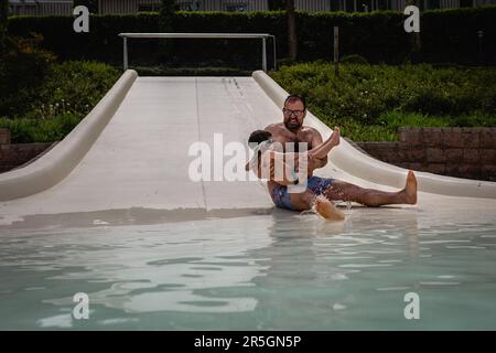 Ein lächelnder Vater hält seine kleine Tochter in den Händen und rutscht auf der Wasserrutsche hinunter, um Spaß zu haben Stockfoto