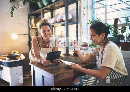 Wo Wirtschaft und Kunst in perfekter Harmonie nebeneinander existieren. Zwei junge Frauen, die während eines Meetings in einem Töpferstudio ein digitales Tablet benutzen. Stockfoto