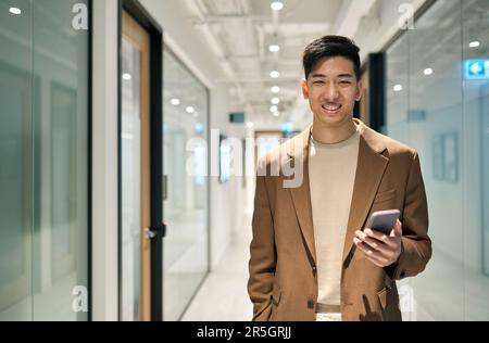 Junger, glücklicher asiatischer Geschäftsmann, der im Flur steht und ein Telefon hält. Stockfoto