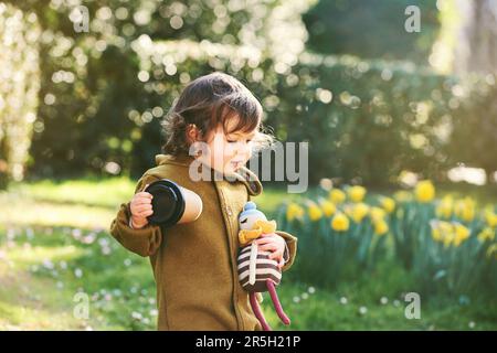 Außenporträt eines niedlichen Kleinkindes, das im Spring Park spielt, eine Tasse Kaffee zum Mitnehmen hält und eine Strickpuppe hält Stockfoto