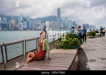 Eine attraktive junge Chinesin posiert für Ein Foto auf der Avenue of Stars mit Hong Kong Island als Kulisse, Kowloon, Hongkong, China. Stockfoto