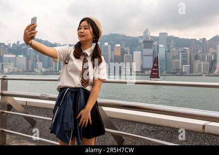 Eine junge Chinesin nimmt Ein Selfie auf der Avenue of Stars mit Hong Kong Island als Kulisse, Kowloon, Hongkong, China. Stockfoto