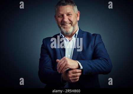 Man muss die Rolle spielen, um die Rolle zu spielen. Studioporträt eines selbstbewussten und reifen Geschäftsmanns, der seine Handschellen vor einem dunklen Hintergrund zuknöpft. Stockfoto