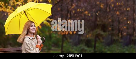 Glückliche Frau mit Regenschirm, die an regnerischen Tagen im Herbstpark auf einer Bank sitzt, Platz für Text. Bannerdesign Stockfoto