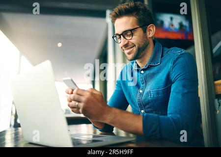 Ich beantworte meine SMS, bevor ich arbeite. Ein gutaussehender junger Geschäftsmann, der im Café an seinem Laptop arbeitet und sein Handy benutzt. Stockfoto