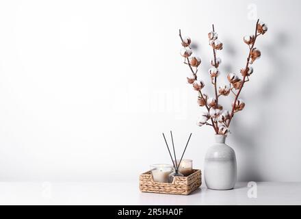 Brennende Kerzen in Korb und Vase mit Baumwollzweigen auf dem Tisch vor weißem Hintergrund. Platz für Text Stockfoto