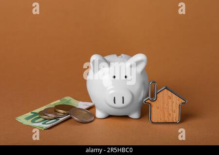 Keramik-Sparschwein, Geld und Schlüsselartikel auf braunem Hintergrund. Finanzielle Einsparungen Stockfoto