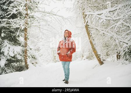 Außenporträt eines Mannes mittleren Alters, der einen schönen Tag im Winterwald genießt, unter Schneefall spaziert, eine rote orangefarbene Jacke trägt Stockfoto