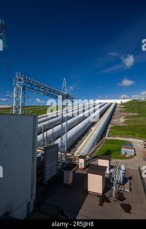 Rohre des Wasserkraftwerks und Transformatorstation mit Elektroturm (links) auf blauem Himmelshintergrund Stockfoto