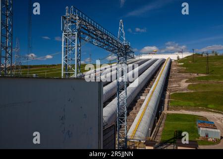 Rohre des Wasserkraftwerks (rechts) und Elektromast auf blauem Himmelshintergrund Stockfoto