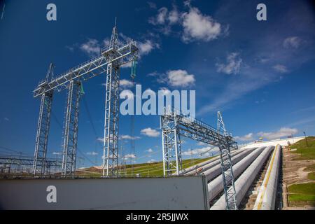 Rohre des Wasserkraftwerks (rechts) und elektrische Türme auf blauem Himmelshintergrund. Stockfoto