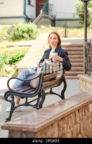 Rothaarige Frau mittleren Alters, die auf einer Bank sitzt und Kaffee und Croissant hält und Spaß hat Stockfoto