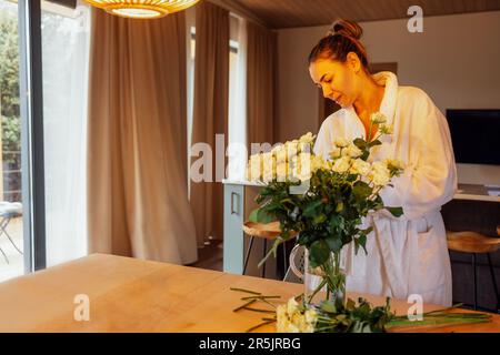 Junge, charmante Frau im weißen Bademantel schneidet Stämme weißer Rosen und steckt sie in eine Glasvase. Lächelndes, attraktives Mädchen im Hotelzimmer oder in der Küche Stockfoto