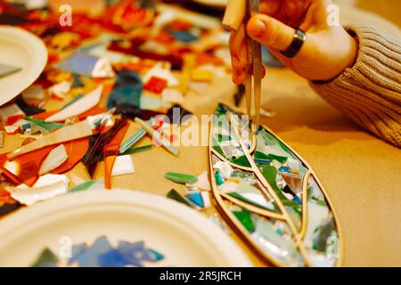 Weibliche Hand mit Ringen, die während der Mosaikherstellung ein Glasstück mit einer Pinzette festhält. Teenager-Mädchen stecken ein grünes Stück Glas in die Form eines Baumblattes. Mehrspaltig Stockfoto