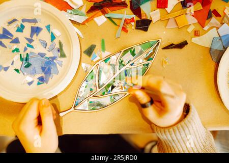 Weibliche Hand mit Ringen, die während der Mosaikherstellung ein Glasstück mit einer Pinzette festhält. Teenager-Mädchen stecken ein grünes Stück Glas in die Form eines Baumblattes. Mehrspaltig Stockfoto