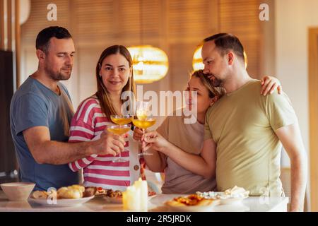 Zwei verheiratete Paare halten ein Glas Champagner und machen Toast im Speisesaal zu Hause. Junge, lächelnde Menschen in lässiger Kleidung knallen Becher mit Glitzer Stockfoto