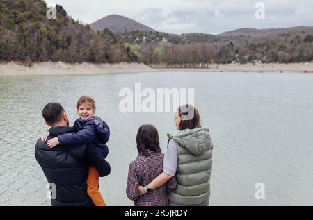 Glückliche Familie mit zwei Kindern am See. Die Leute tragen warme Jacken und Mäntel. Mom, Dad, Teenager und Vorschultochter, die sich zurückhalten. Mann, Frau, Stockfoto